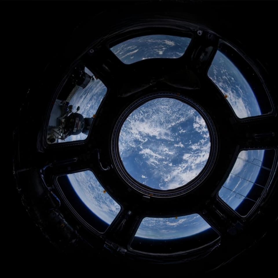 Vista da Terra pelo vitral circular de uma estação espacial, com oceanos e nuvens sobre o planeta.